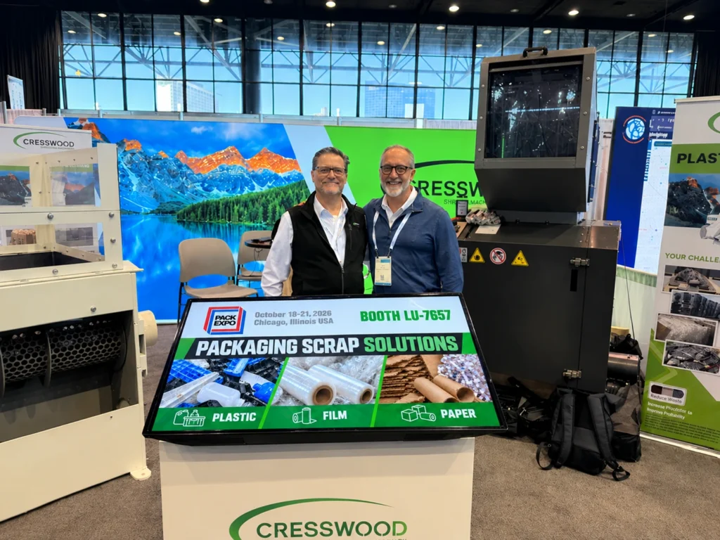 Two men standing in a manufacturing trade show booth surrounded by shredding equipment.